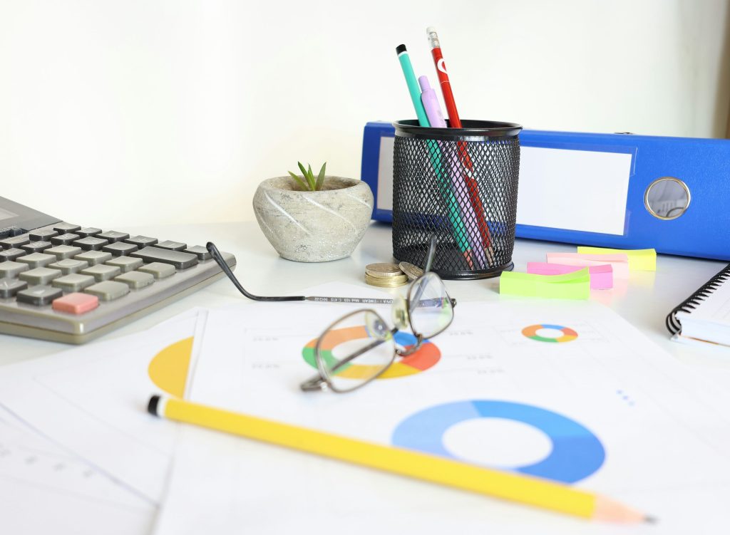 A desk with a calculator, a pair of glasses, printed charts and office supplies